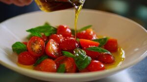 Olive oil being poured onto a salad of halved cherry tomatoes and fresh basil leaves in a white bowl