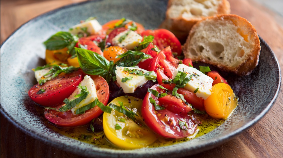 A bowl of heirloom tomato salad with mozzarella cheese, fresh basil, and olive oil, accompanied by slices of crusty bread