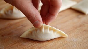 Close-up of a hand folding a wonton wrapper with pleats on a wooden board
