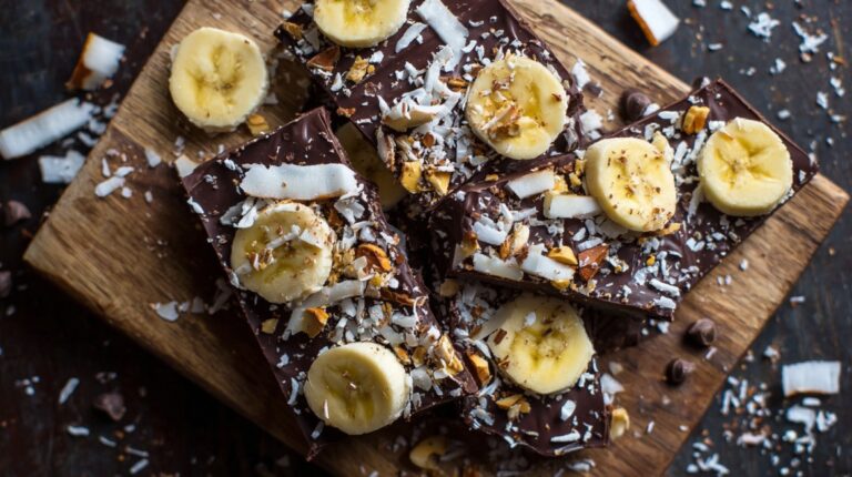 Overhead view of chocolate bark topped with banana slices, shredded coconut, and chopped nuts on a wooden board
