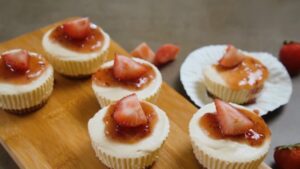 Strawberry cheesecake bites topped with strawberry sauce and fresh strawberry slices are arranged on a wooden board