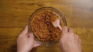 A person mixes a crumbly cheesecake base mixture in a glass bowl using a spatula