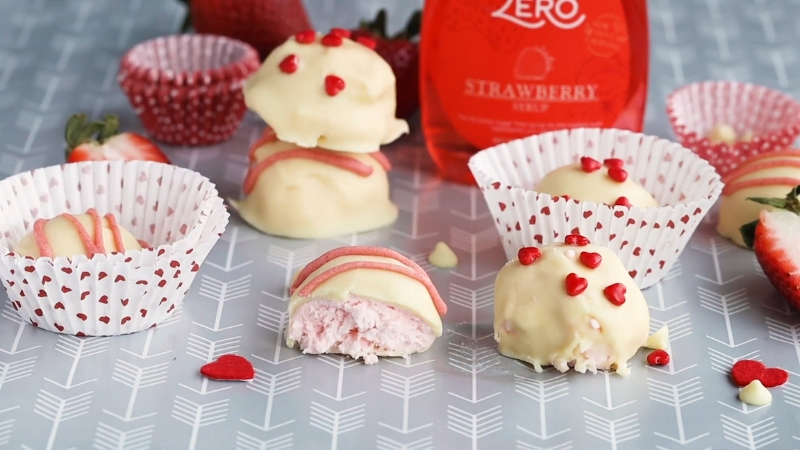 White chocolate-covered strawberry cheesecake bites decorated with red heart sprinkles and pink drizzle, some in cupcake liners and one cut open to show the filling