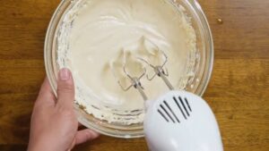 A person uses a hand mixer to blend creamy cheesecake filling in a glass bowl