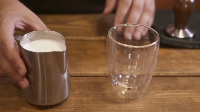 A person holds a metal pitcher of frothed pea milk beside an empty glass on a wooden counter
