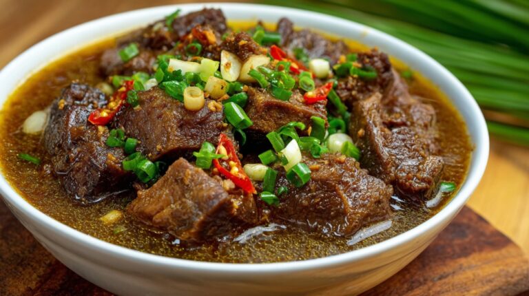 Bowl of braised beef pieces in dark broth topped with scallions and chili.